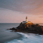 Sunset illuminates a lighthouse perched on a rocky shoreline, creating a serene seascape.